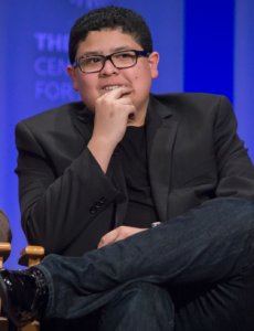 Rico Rodriguez seated looking puzzled in a black jacket and wearing glasses